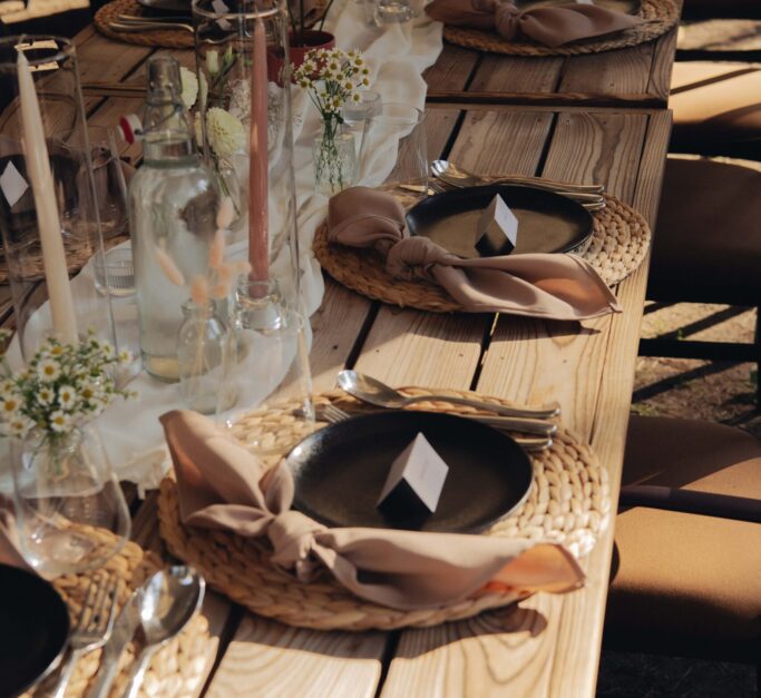 Elegantiškai išklotas stalas lauko vakarienei su rudos spalvos detalemis ir šviesiomis gėlėmis.