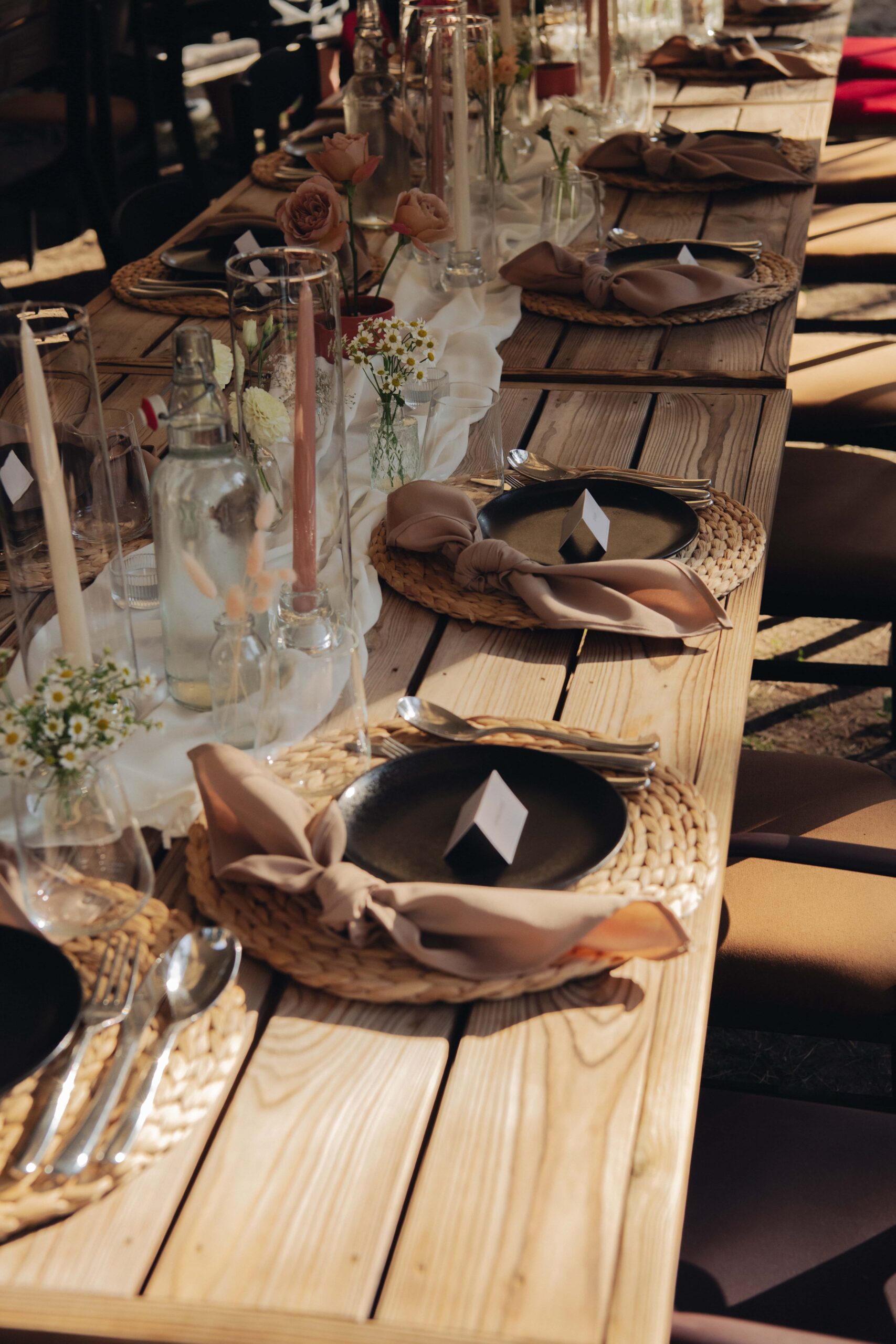 Elegantiškai išklotas stalas lauko vakarienei su rudos spalvos detalemis ir šviesiomis gėlėmis.